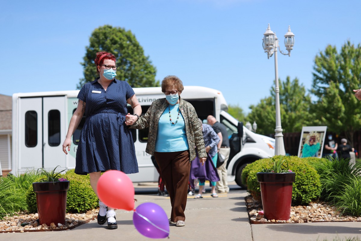 A caregiver and an elderly person walk together outdoors, both wearing masks, with colorful balloons nearby.