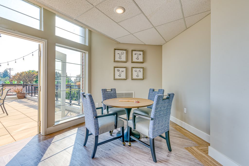 A small meeting room with a round table and four chairs, featuring large windows and framed artwork on the wall.