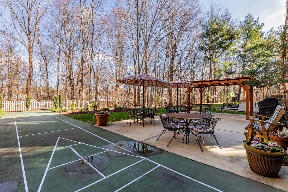 A backyard patio with a pergola and seating area next to a shuffleboard court surrounded by trees.