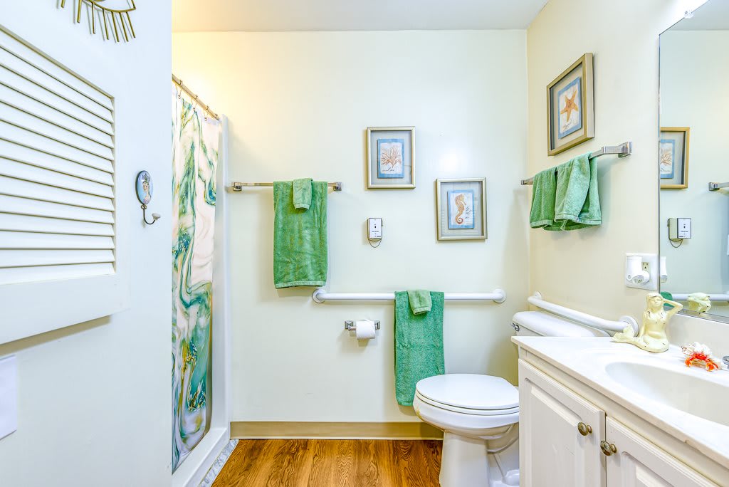 A clean and well-organized bathroom featuring green towels, framed wall art, a toilet, and a shower with a decorative curtain.