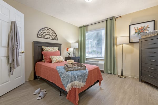 A cozy bedroom featuring a single bed adorned with decorative pillows and a colorful blanket, complemented by a lamp and window with green curtains.