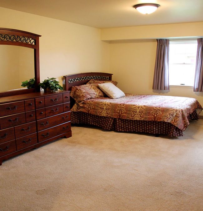 A spacious bedroom featuring a bed with pillows, a dresser with a mirror, carpet flooring, and curtains on the window.