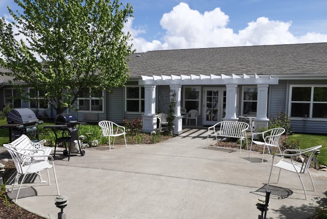 A sunny day view of a house with a patio, outdoor chairs, and a tree in the yard.