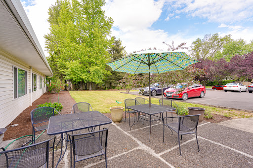 A cozy outdoor seating area with tables and umbrellas, surrounded by lush greenery and a parking lot.