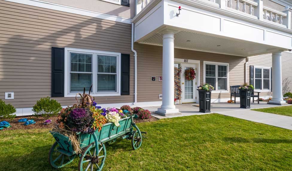 A well-maintained building entrance with a decorative wheelbarrow full of flowers and a lush green lawn.