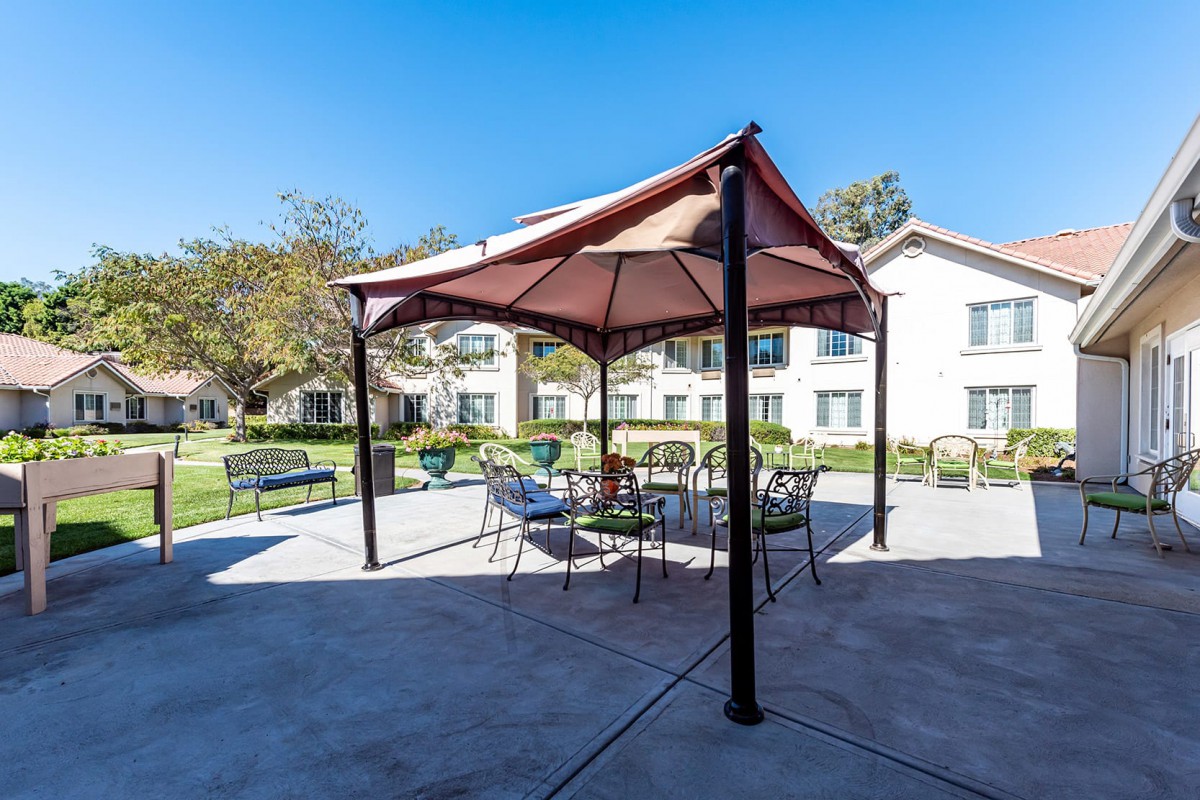 A sunny outdoor area featuring a gazebo with patio furniture, surrounded by benches and greenery, with buildings in the background.