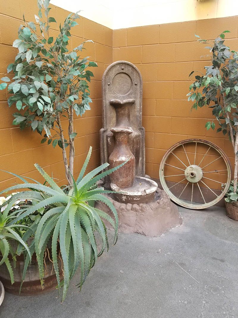 A rustic outdoor setup featuring a decorative fountain, potted plants, and an old wagon wheel against a warm-colored wall.