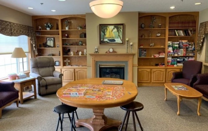 A cozy living room featuring a round table with a puzzle in progress, surrounded by bookshelves and an armchair near a fireplace.
