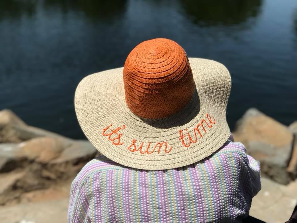 A person wearing a sun hat with the phrase 'it's sun time' embroidered on it, overlooking a lake.