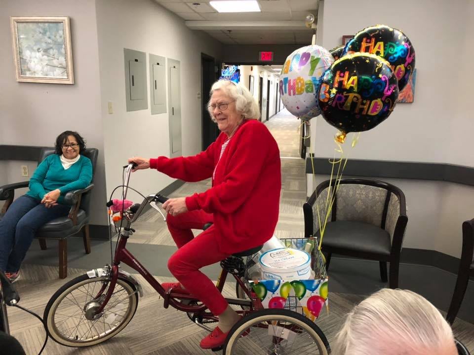 An elderly person in red clothing rides a bicycle indoors with celebration balloons attached.