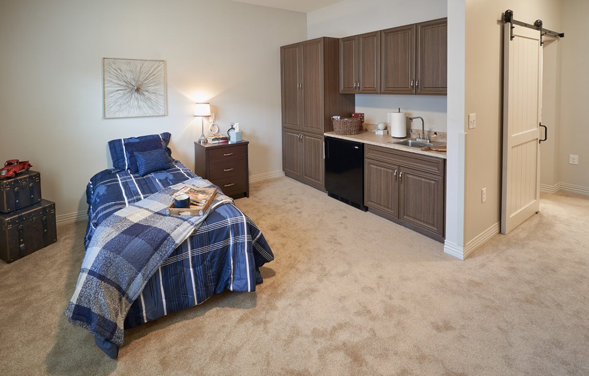 A cozy bedroom with a small kitchenette, featuring a bed with blue plaid bedding and wooden furniture.