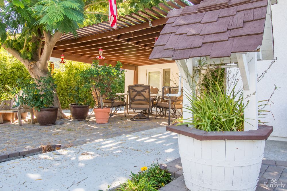 A cozy patio area with chairs under a wooden pergola, surrounded by lush potted plants.