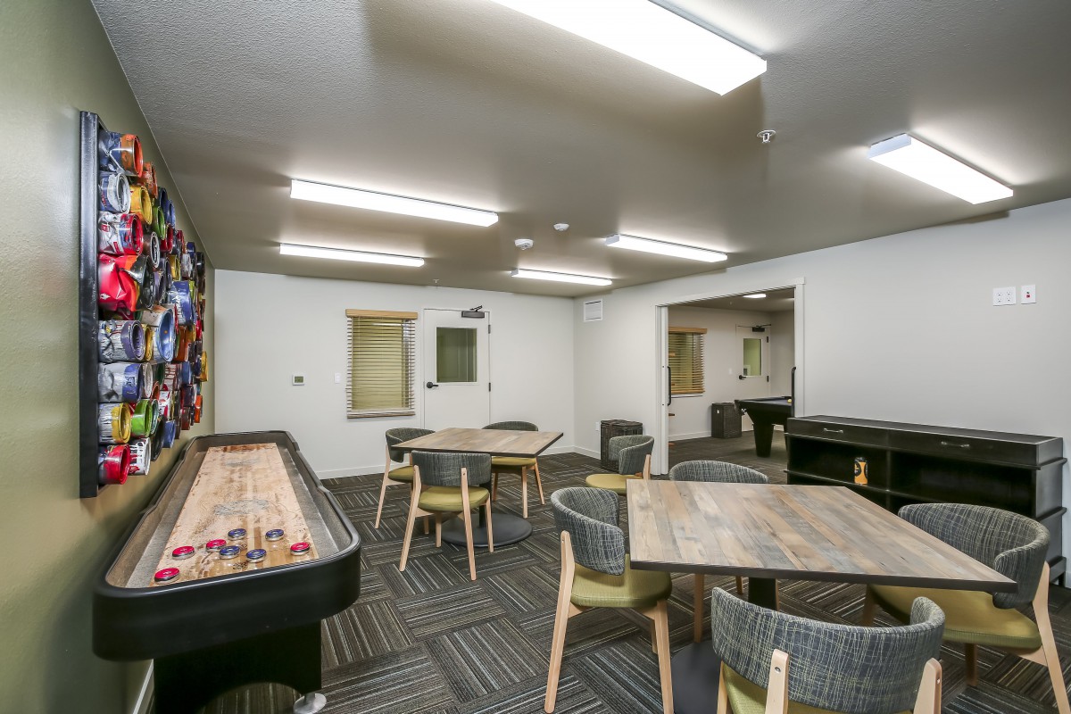 A well-lit indoor game room featuring a shuffleboard table, several wooden tables and chairs, creating a comfortable recreational space.