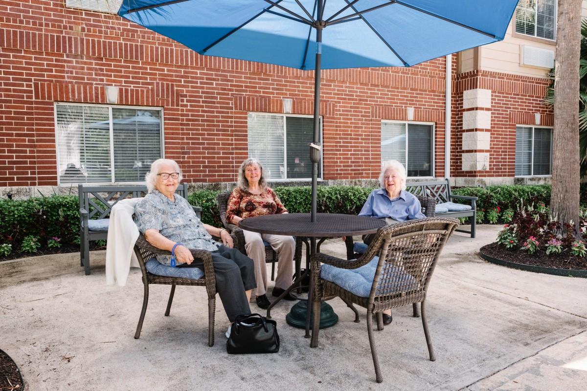 Three seniors sit around a table under a blue umbrella on a patio.