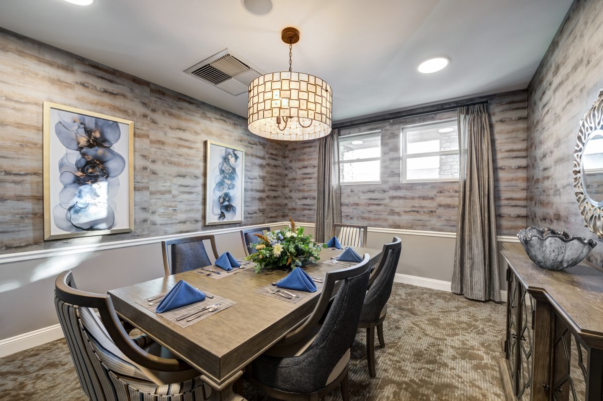 A stylish dining room with a rectangular table set for six, adorned with blue napkins and a central floral arrangement. The room features elegant chairs, a modern chandelier, decorative wall art, and grey curtains complementing the overall decor.