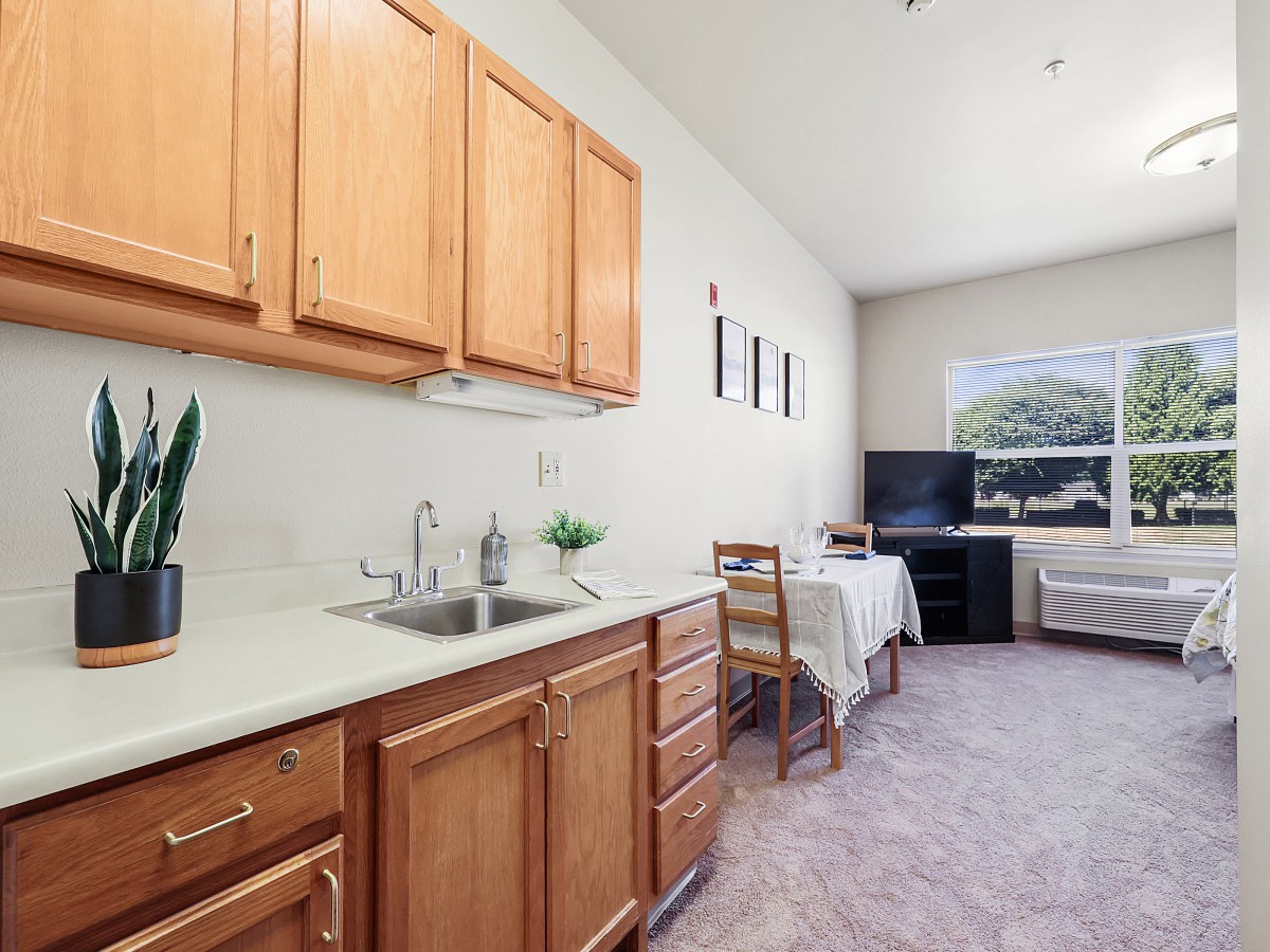 A compact kitchen area with wooden cabinets and a sink, leading to a small dining space with a table, chairs, and a television.