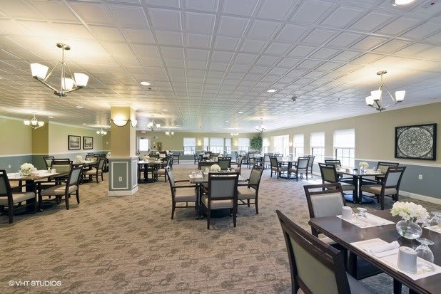 A spacious and well-lit dining area with multiple tables and chairs arranged on a carpeted floor, surrounded by large windows.