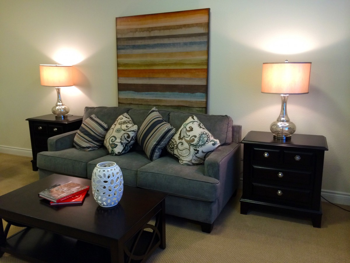 A cozy living room featuring a gray sofa with patterned throw pillows, flanked by matching table lamps and side tables. An abstract painting hangs above the sofa.