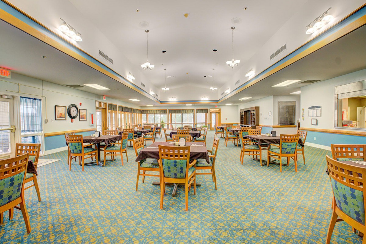 A spacious dining room with several tables and chairs, featuring a patterned carpet and overhead lighting.