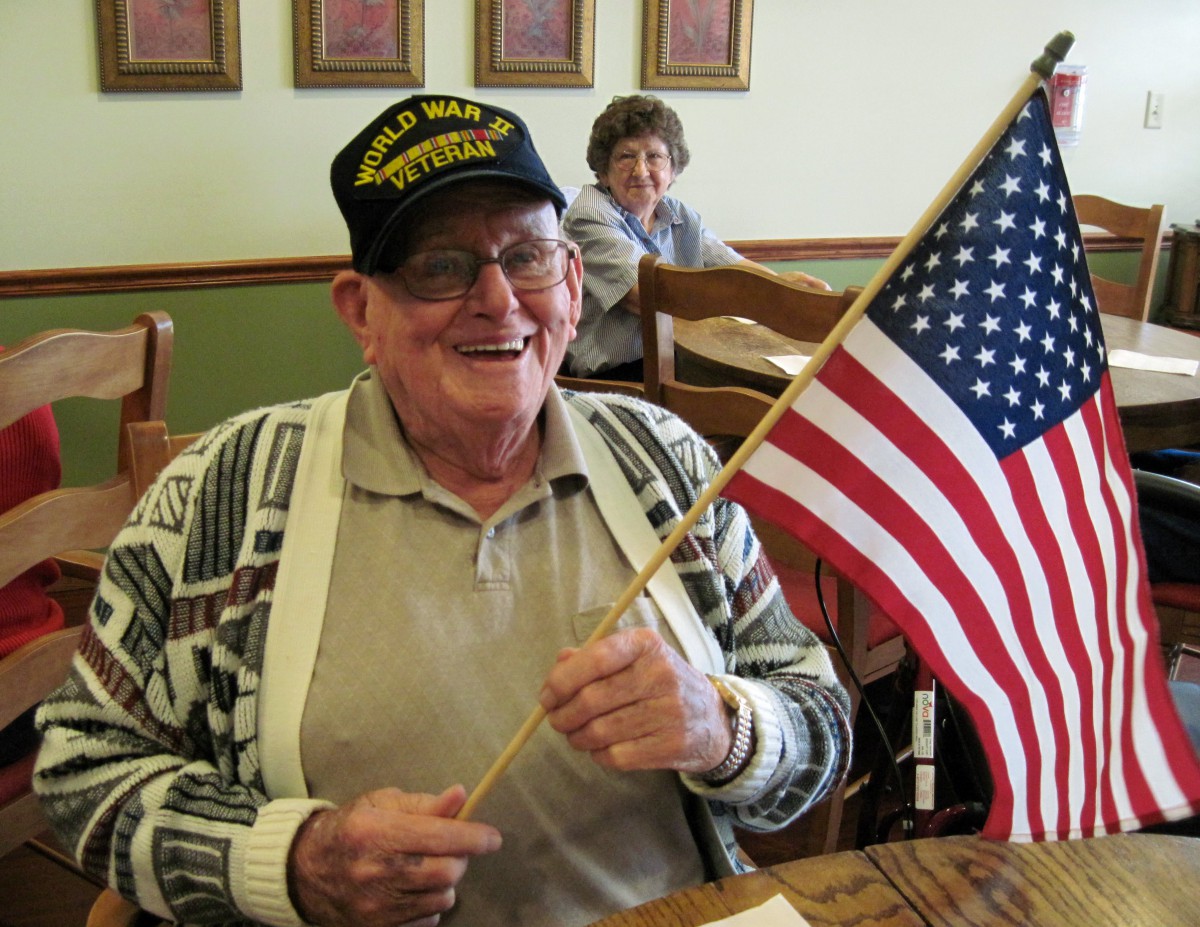An elderly man smiling while holding an American flag indoors.