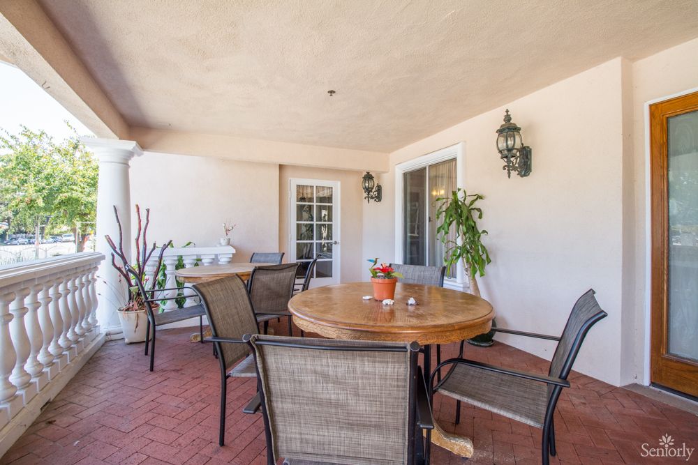 A cozy patio area with a round wooden table, surrounded by chairs and decorated with plants and wall lanterns.