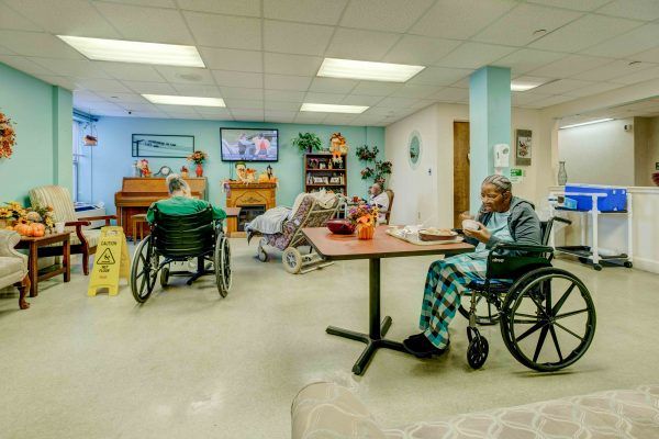 A common area in a nursing home with elderly residents seated in wheelchairs, engaging in various activities.