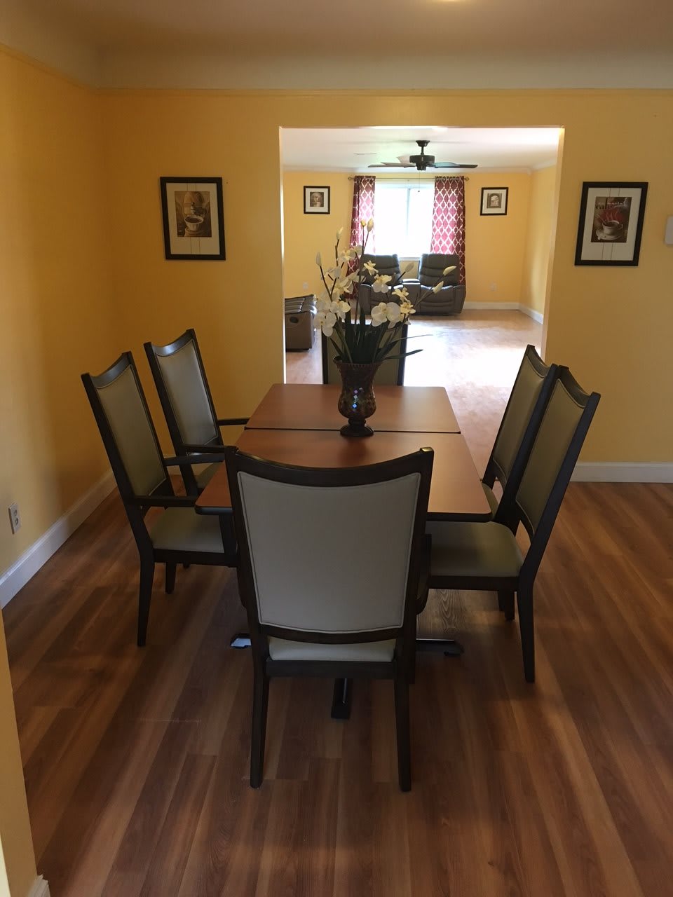 A neatly arranged dining room with a wooden table and six chairs, surrounded by yellow walls and decorated with framed art.