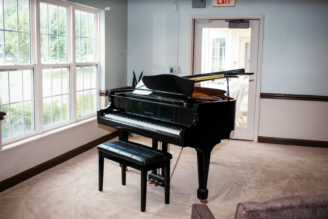 A grand piano sits in a well-lit room with large windows and a bench.
