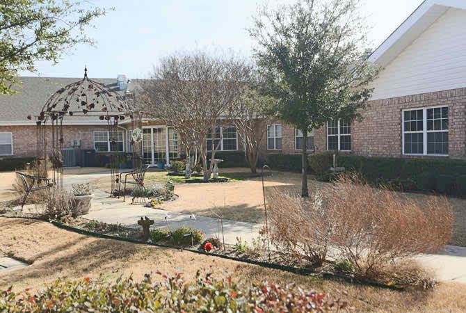 A serene garden with a circular pathway surrounded by trees and plants, adjacent to a building.