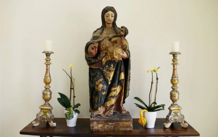 A wooden statue of the Virgin Mary holding baby Jesus, flanked by two ornate candles and potted orchids on an altar.