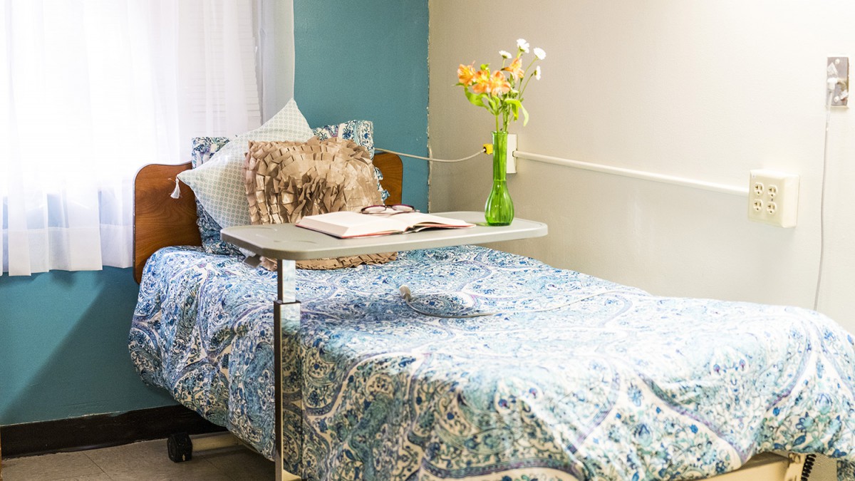A cozy hospital room with a bed, decorative pillows, and fresh flowers in a vase on a tray table.