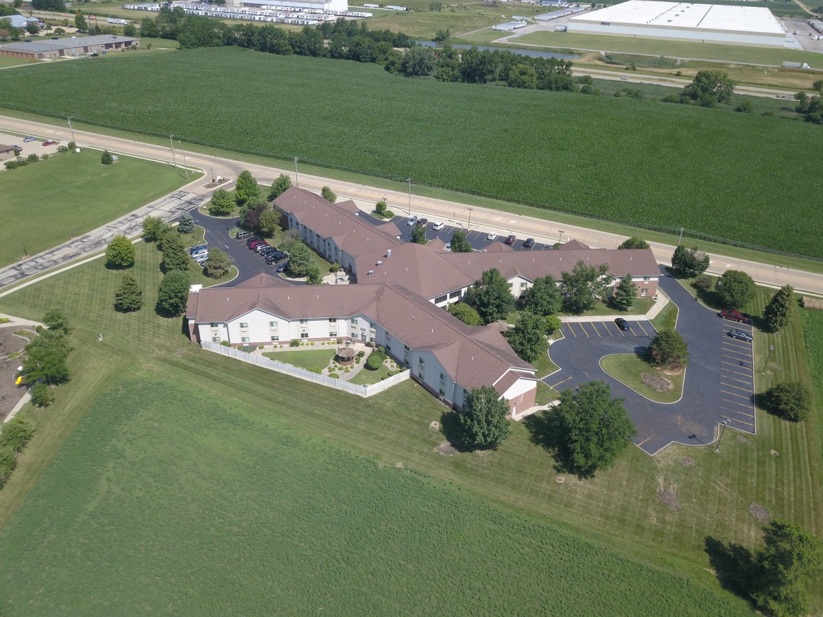 Aerial view of a large building surrounded by green fields and trees, with a parking area adjacent.