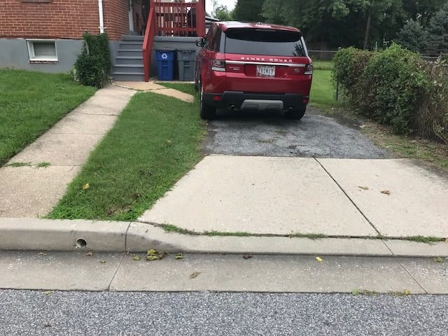 A red SUV parked in a driveway next to a sidewalk and lawn in front of a brick house.