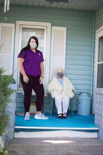A caregiver stands beside an elderly person on a porch, both wearing masks.