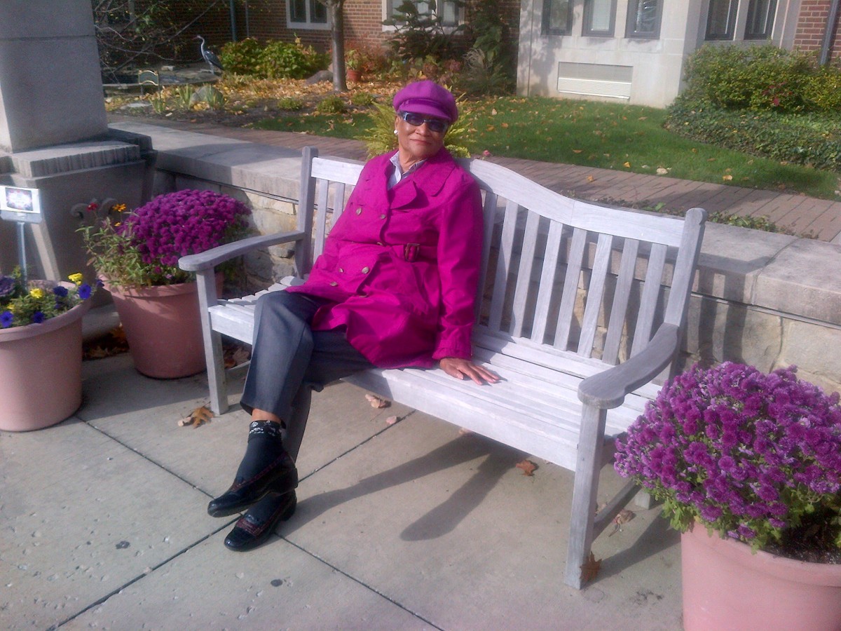 A person sitting on a bench wearing a purple coat and hat, surrounded by potted flowers.
