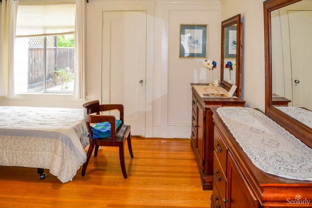 A cozy bedroom with wooden flooring, featuring a bed, dresser with mirror, and a chair by the window.
