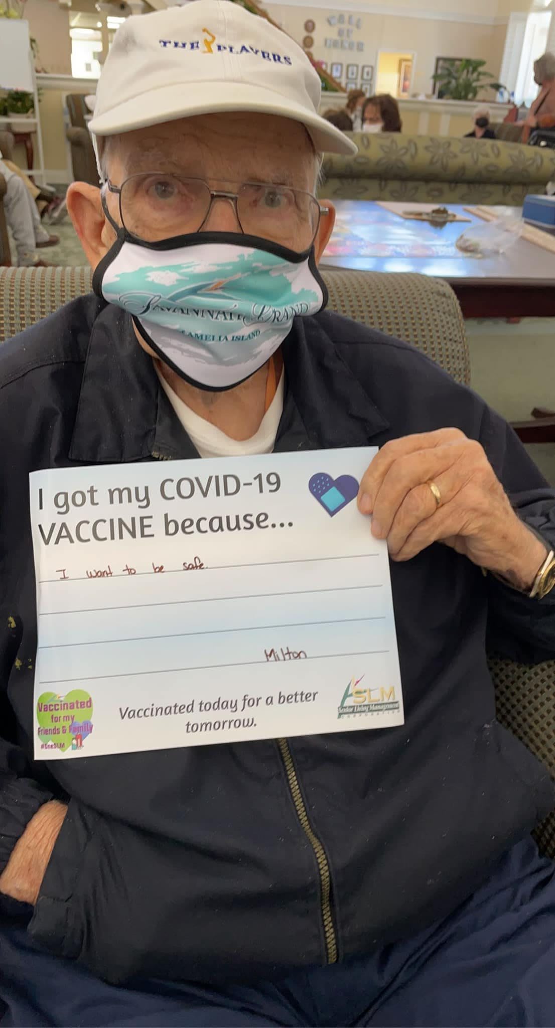 An elderly person wearing a mask holds a sign explaining their reason for receiving the COVID-19 vaccine.