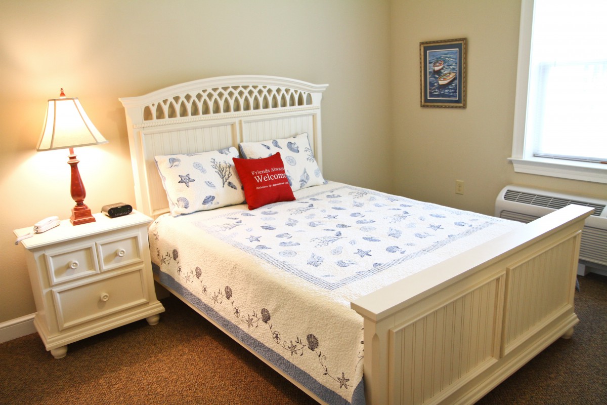 A cozy bedroom with a neatly made bed featuring a white quilt and decorative pillows, accompanied by a nightstand and lamp.