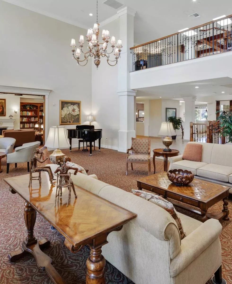 A spacious and elegantly furnished living room with a chandelier, featuring sofas, coffee tables, and a grand piano.