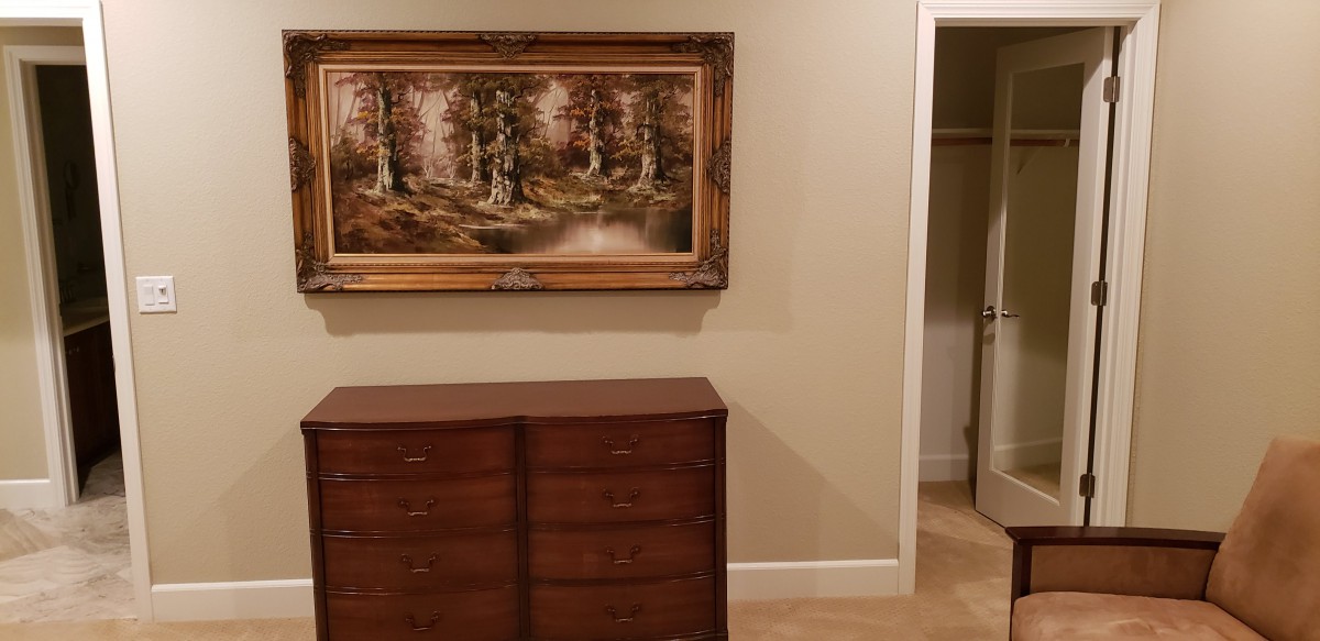 A bedroom interior featuring a wooden dresser, a framed painting of a forest scene, and an open closet with neutral-colored walls.