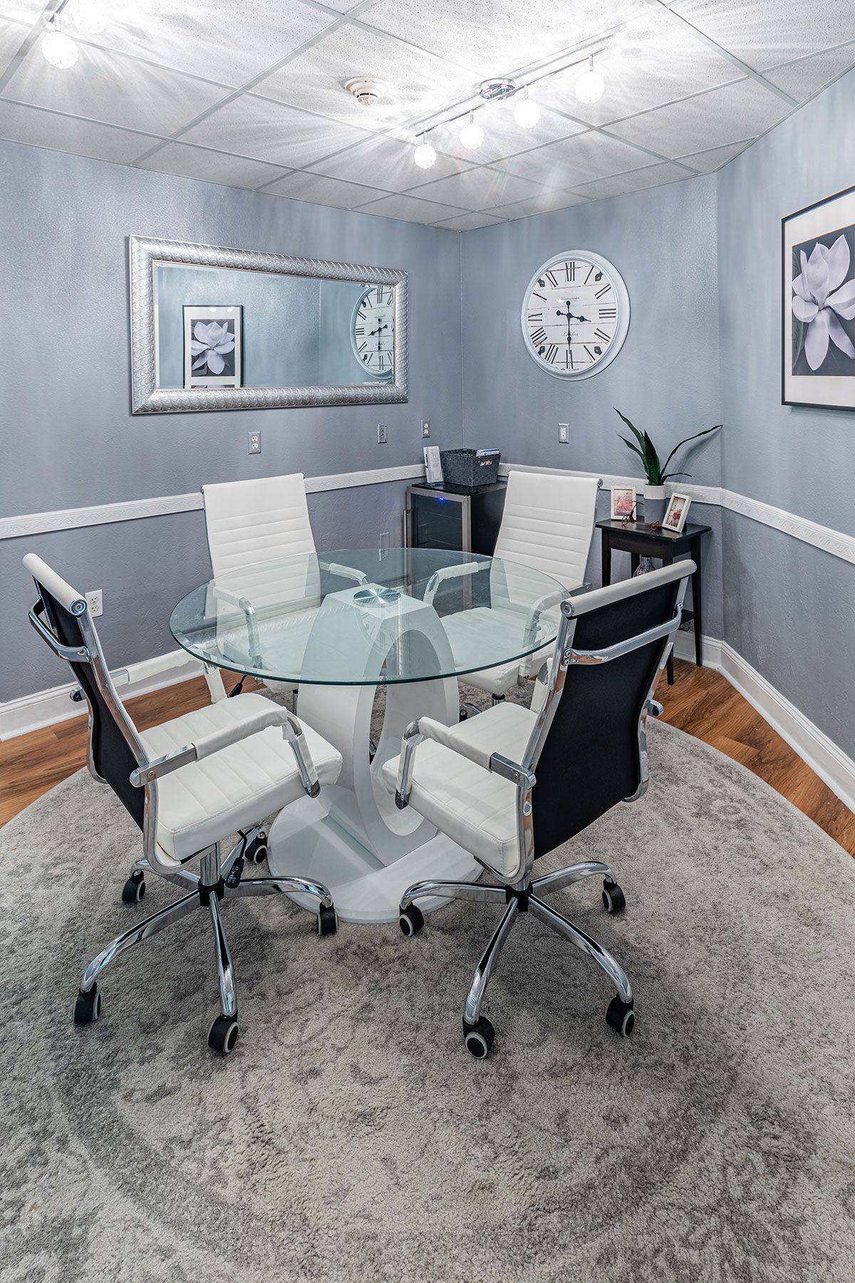 A modern office conference room with a glass table and four chairs, decorated with wall art and a large clock.