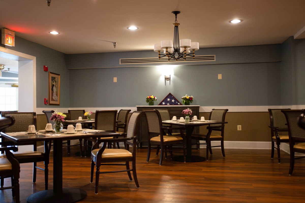 A cozy dining room with neatly arranged tables and chairs, featuring a chandelier and wooden flooring.
