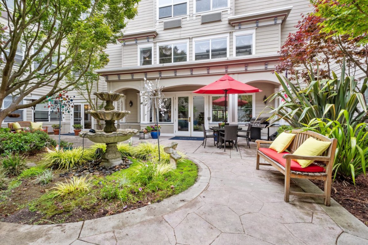 A charming outdoor patio with a fountain, surrounded by lush greenery and comfortable seating under a red umbrella.
