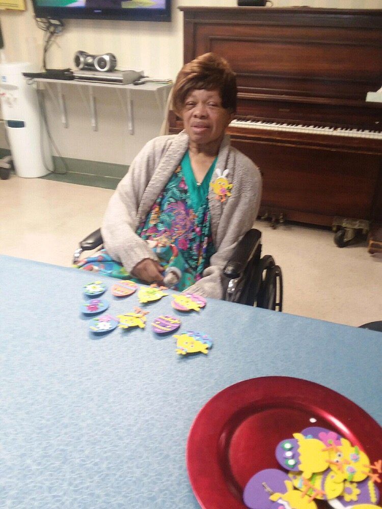 An elderly woman sitting in a wheelchair indoors, with decorations on the table and a piano in the background.