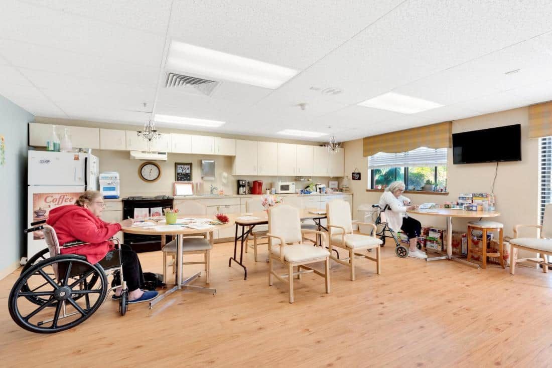 A bright and spacious community room in a nursing home, featuring tables, chairs, and residents in wheelchairs.