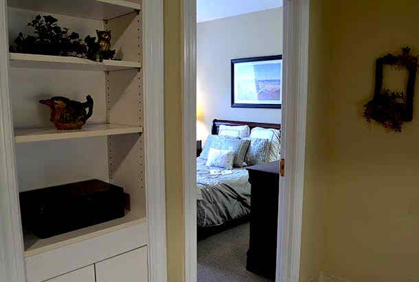 A view into a cozy bedroom through an open doorway, with a neatly arranged bed and a built-in shelf on the side.