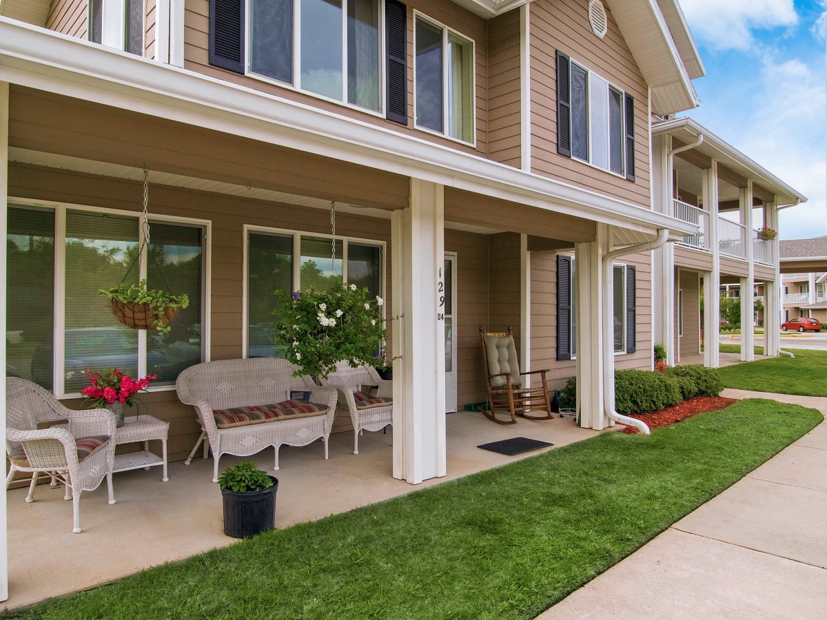 A view of a residential apartment building with a porch area adorned with furniture and plants, surrounded by a well-manicured lawn.