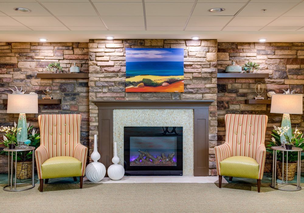 A cozy living room with a stone fireplace, two chairs, and colorful artwork above.