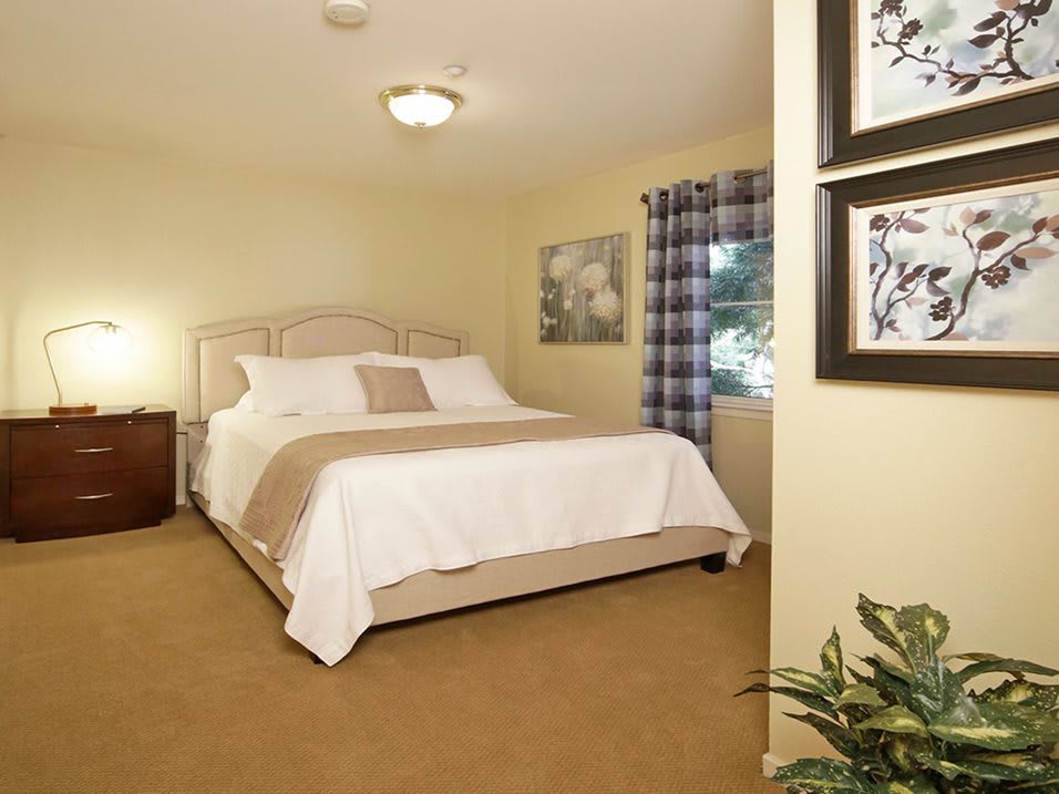 A cozy bedroom with a neatly made bed, framed art on the walls, and a window with plaid curtains.