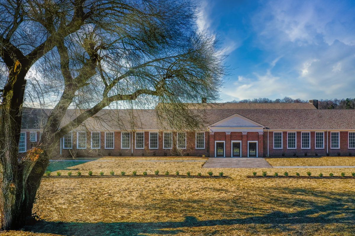 A large brick building with a symmetrical facade and a sloped roof, set against a clear blue sky, surrounded by trees and open lawns.